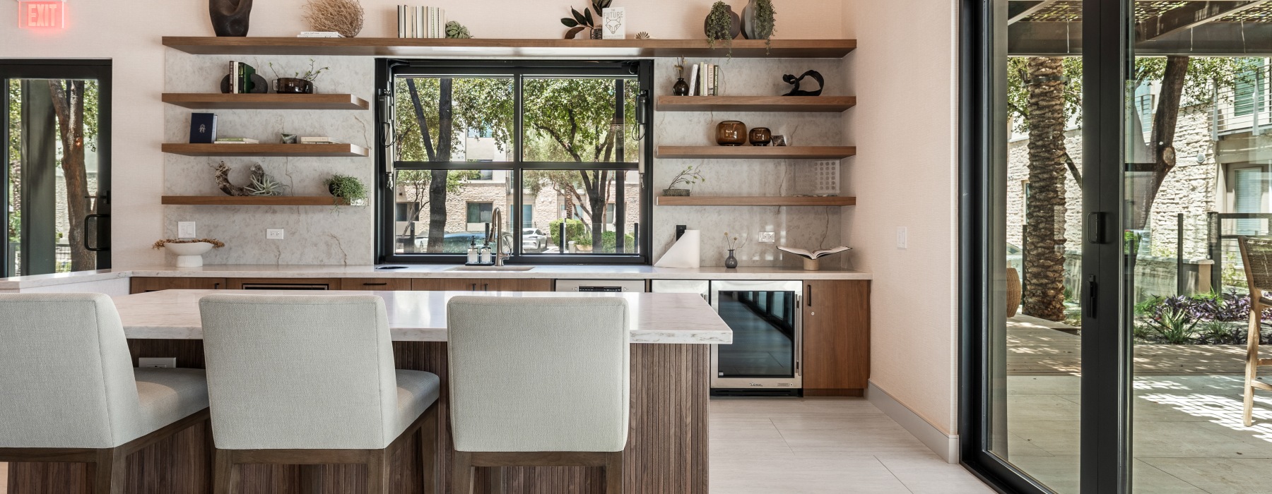 Golf lounge wet bar with marble countertops, walnut open shelving with plants and décor, white barstools, and sliding glass doors opening to an outdoor courtyard.